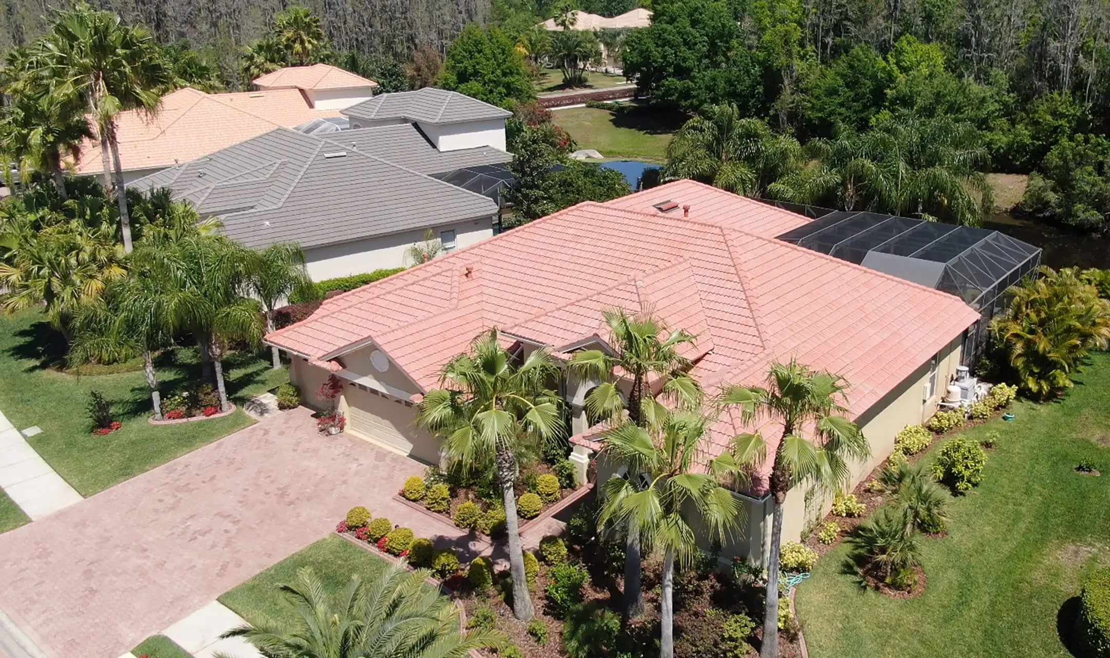 Aerial view of a single-story house with a red-tiled roof, palm trees, landscaped yard, and neighboring homes, set in a green residential area.