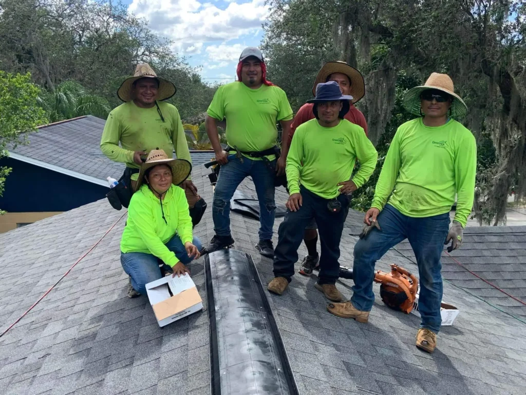 Six construction workers wearing neon green shirts and hats pose and smile on a shingled rooftop under a partly cloudy sky, surrounded by trees. Some hold tools and equipment, and one person is kneeling near a box.