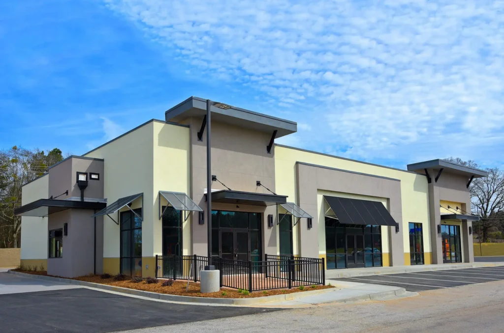 A newly constructed, modern beige and gray commercial building with black awnings and large glass windows, situated in an empty parking lot under a partly cloudy blue sky.