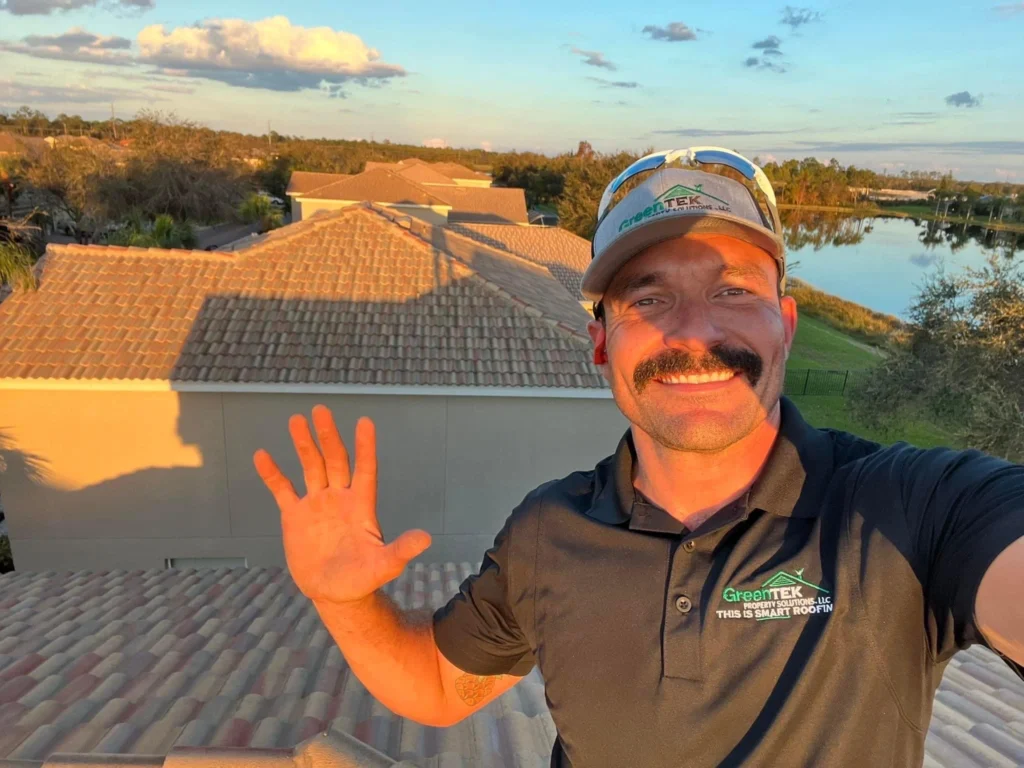 A smiling man with a mustache waves while standing on a tiled roof at sunset. He wears a GreenTEK Roofing polo and cap. Houses, a lake, and trees are visible in the background under a blue sky with clouds.