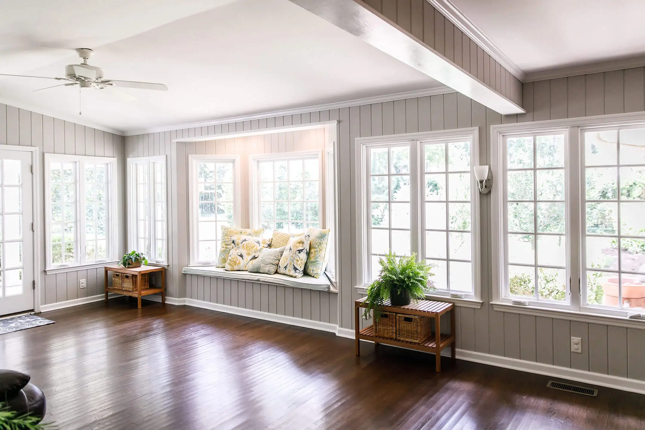 Bright sunroom with large windows, gray paneled walls, dark wood floors, a ceiling fan, a cushioned window seat with floral pillows, and small wooden tables with potted plants.