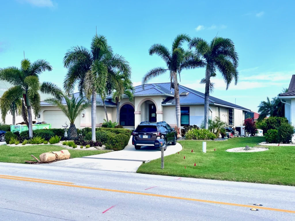 A black car is parked in the driveway of a modern, single-story house with solar panels on the roof, surrounded by palm trees and a well-maintained lawn under a clear blue sky.