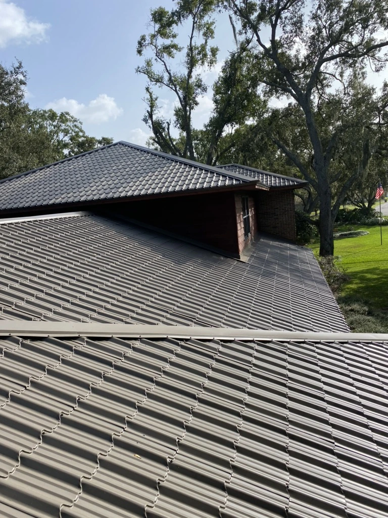 View of a house with a roof made of gray, corrugated metal panels, surrounded by trees and greenery on a sunny day. The sky is mostly clear with a few small clouds.