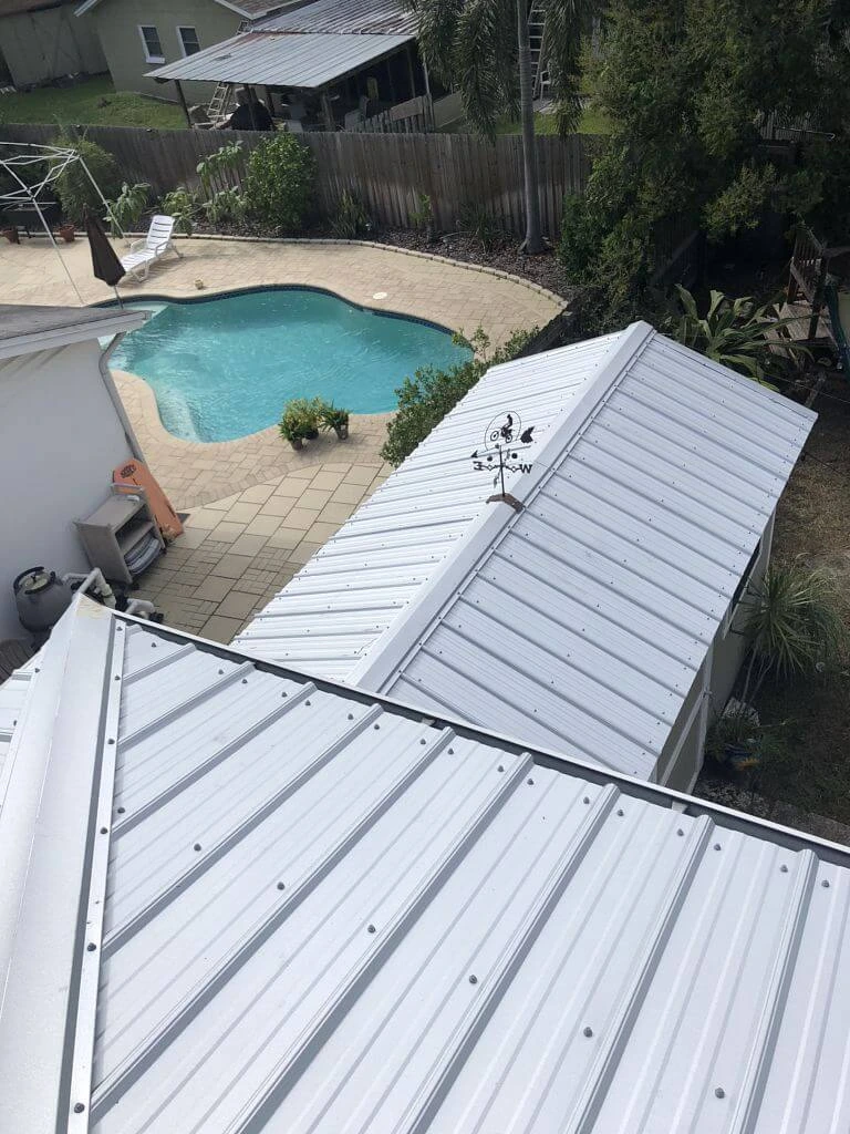 A view from a metal rooftop looking down at a backyard with a swimming pool, patio, wooden fence, and surrounding greenery.