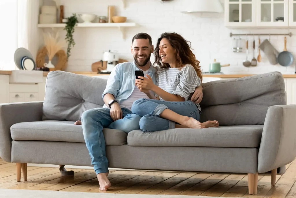 A smiling couple sits close together on a gray couch in a cozy, bright living room, looking at a smartphone and laughing. The background shows a tidy kitchen with shelves and light decor.