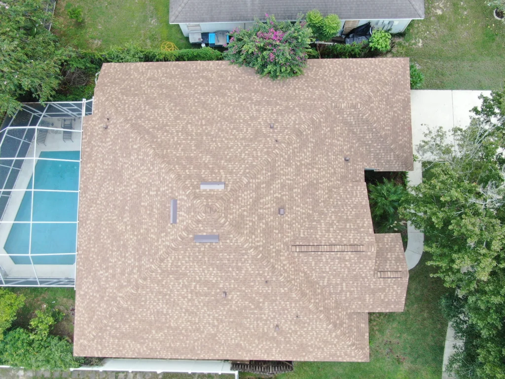 Aerial view of a house with a brown shingle roof, surrounded by green trees and grass. Next to the house is a screened-in swimming pool, and a driveway is partially visible on the right side.