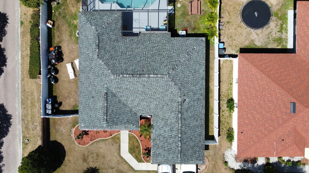 Aerial view of a house with a gray shingle roof, a backyard pool with screened enclosure, a trampoline, vehicles in the driveway, and landscaped yard, surrounded by neighboring homes and fences.