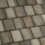 Close-up view of a tiled roof with large, rectangular, overlapping shingles in shades of gray and brown, displaying weathered and slightly varied coloration.