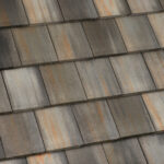 Close-up view of weathered, multicolored roof shingles with shades of gray, brown, and rust, arranged in a neat, overlapping pattern.