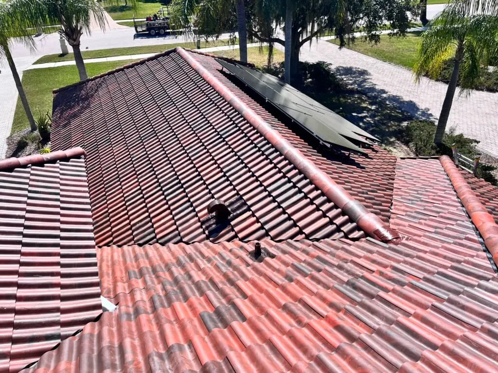 A person is installing solar panels on the red tile roof of a house, with trees, a driveway, and a truck visible in the background on a sunny day.