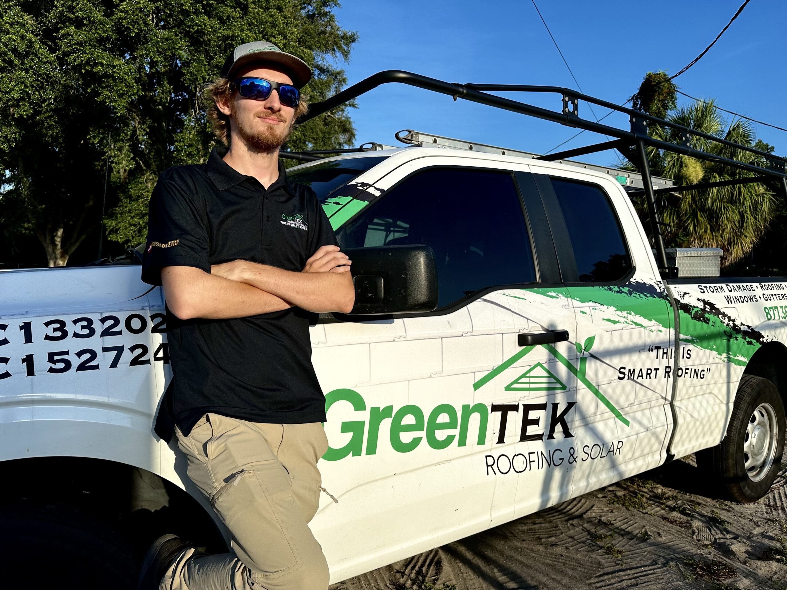A man wearing sunglasses, a cap, and a dark shirt stands with arms crossed in front of a white GreenTEK Roofing & Solar truck in a sunny, outdoor setting with trees in the background.