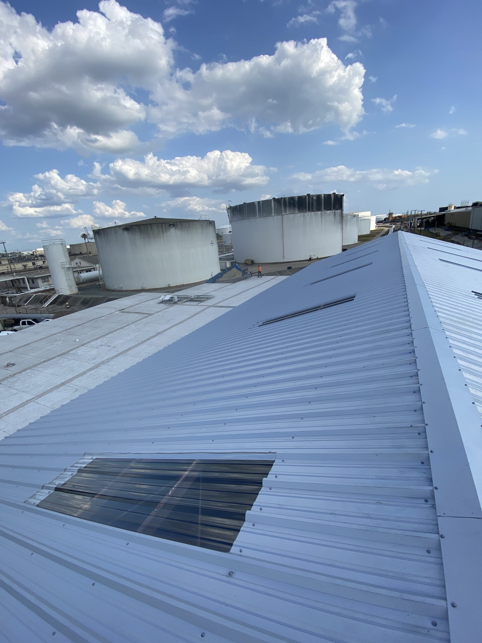 A view from the top of a metal industrial roof with several large cylindrical storage tanks in the background under a blue sky with scattered clouds.