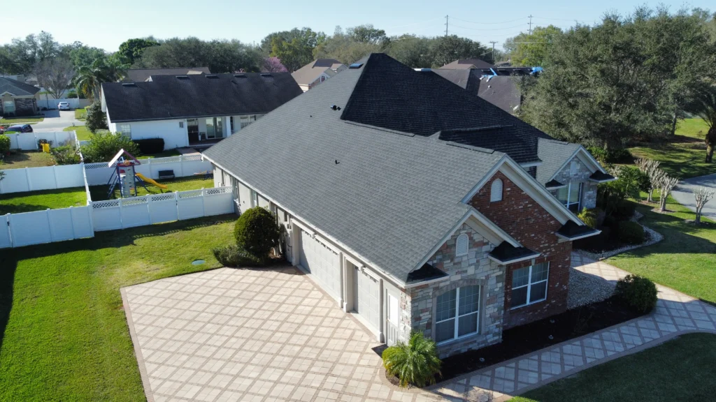 Aerial view of a single-story house with a dark roof, brick and stone exterior, three-car garage, and large paved driveway, surrounded by neatly maintained lawns and white fencing.