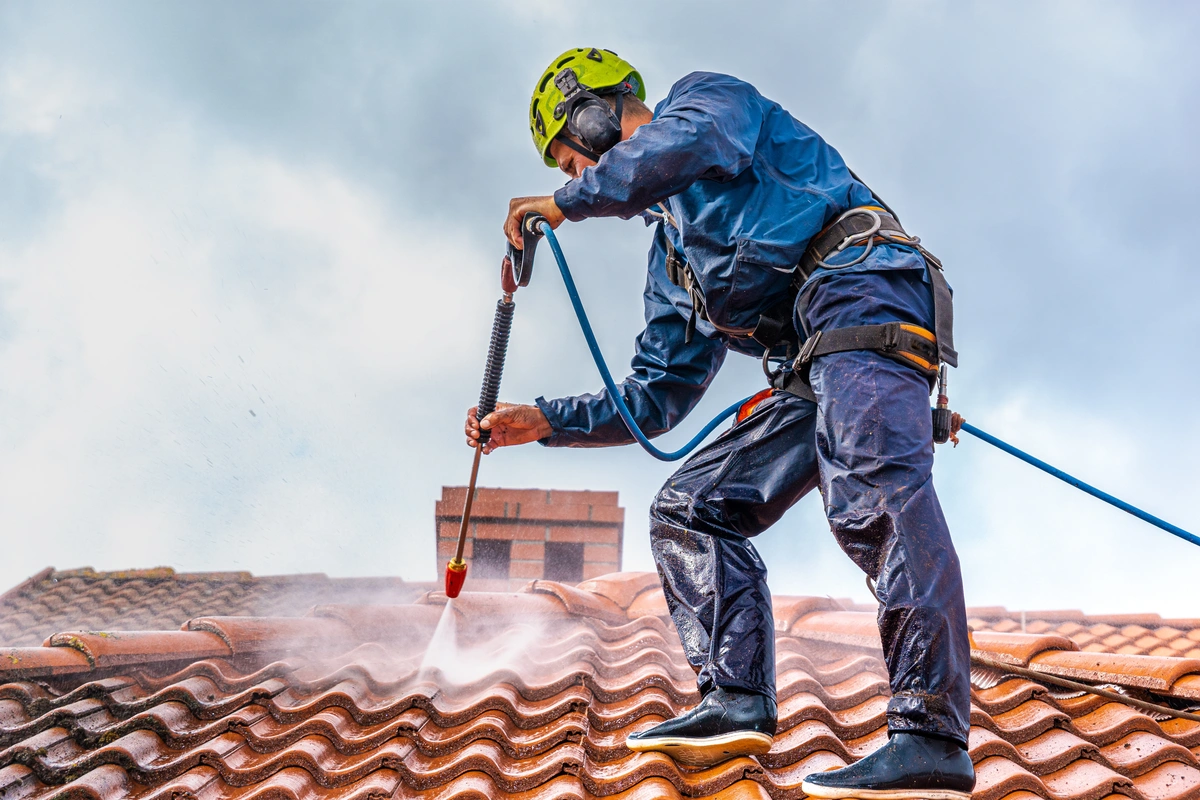 A worker wearing safety gear and a helmet pressure washes a tiled roof, demonstrating the best way to clean a tile roof in Florida as clouds of water spray rise beneath a cloudy sky.