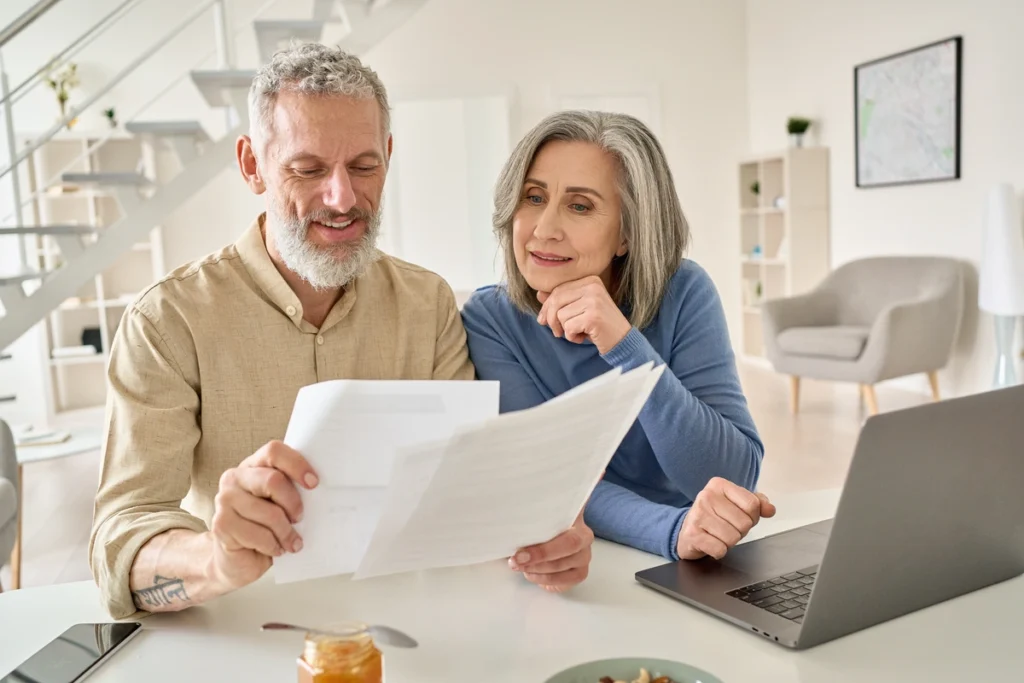 An older couple sits at a table, reviewing roofing Sarasota County documents together and smiling. A laptop is open in front of them, and they appear to be in a bright, modern living room.