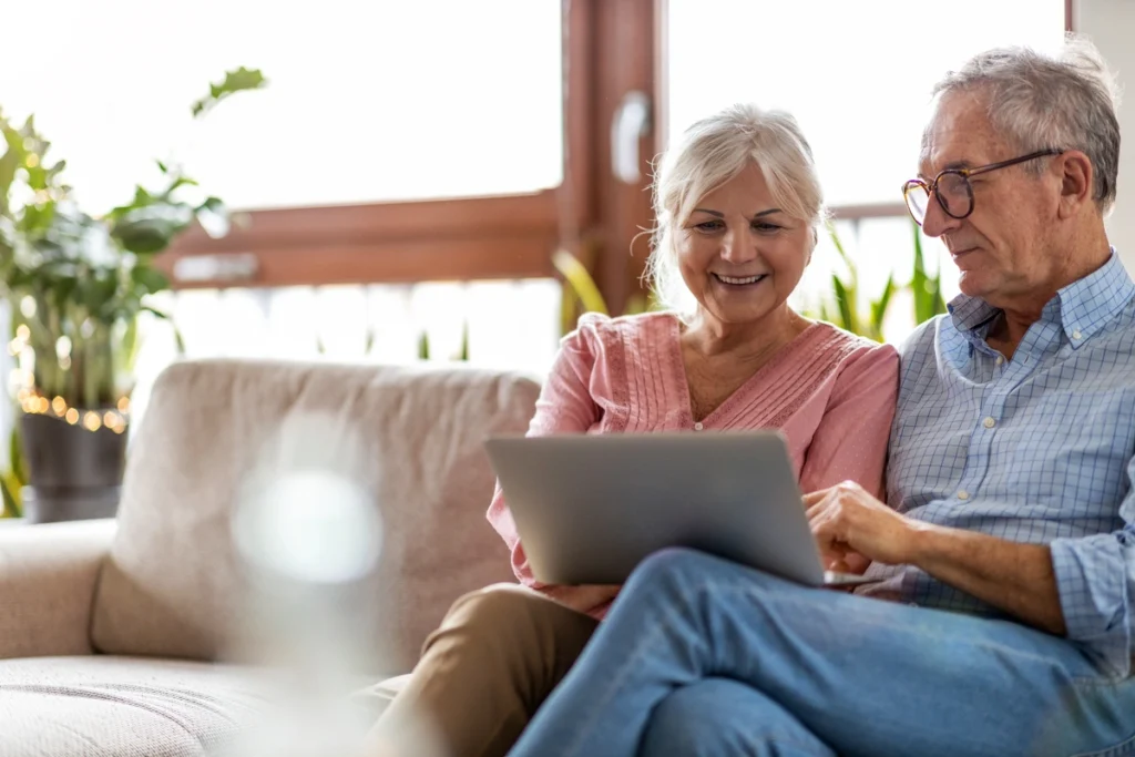 An older couple sits together on a couch, smiling as they look at a laptop, perhaps researching roofing in Polk County. The background features large windows and indoor plants, creating a cozy, well-lit living room atmosphere.