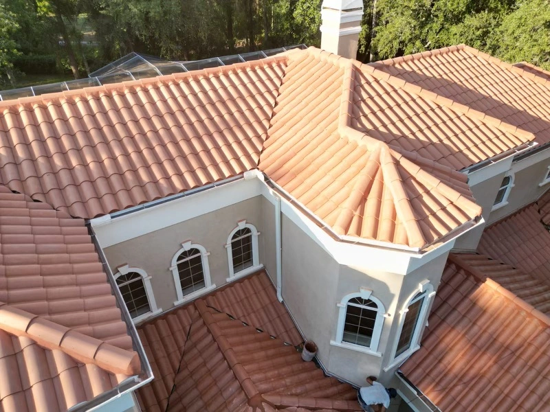 Aerial view of a large house with cream-colored walls and a red clay tile roof featuring multiple slopes, ridges, and arched windows, surrounded by green trees.