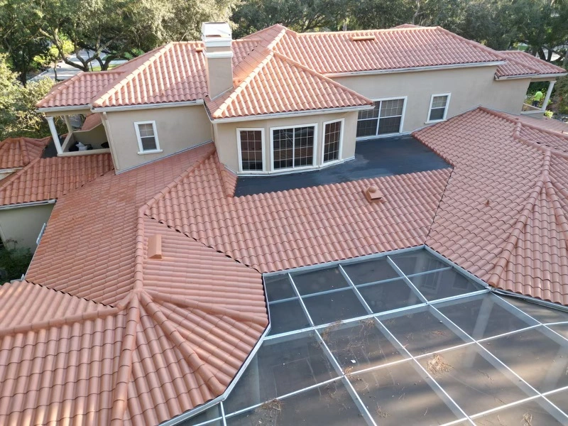 Aerial view of a large house with multiple red tile roofs, beige exterior walls, and a screened patio area. Trees surround the property. Some roof sections appear newer than others.