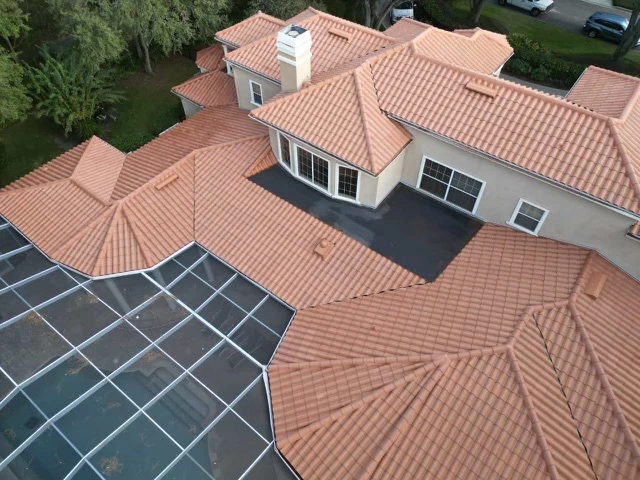 Aerial view of a large house with multiple sections of orange clay tile roofing, a spacious black patio area, and a screened enclosure adjacent to the house, surrounded by trees and greenery.