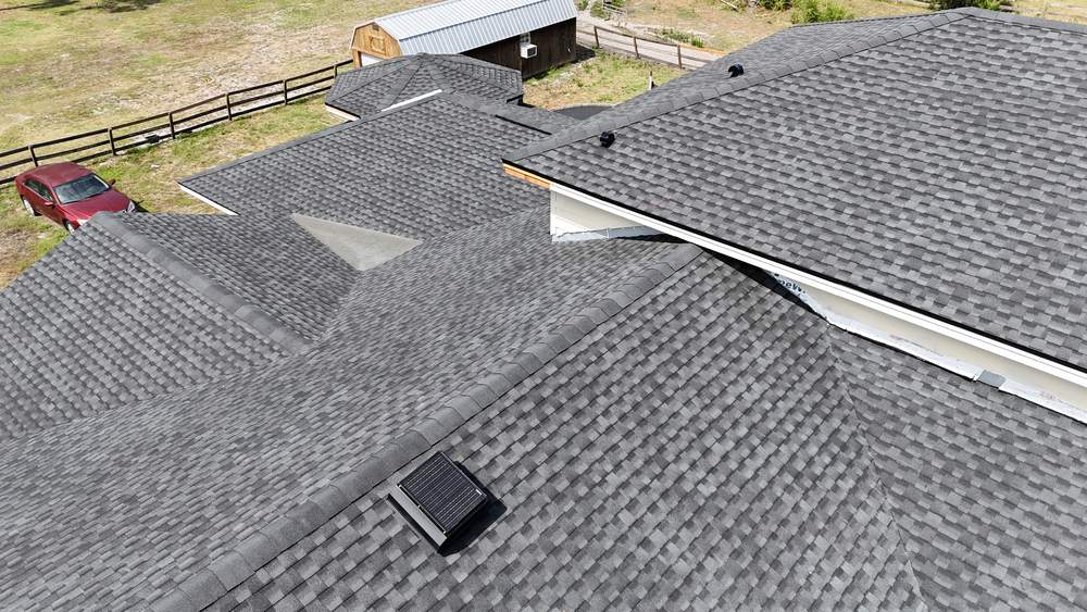 Aerial view of a house with multiple gray shingled roof sections, a solar attic vent, a nearby red vehicle, a wooden fence, and a small shed in the background.