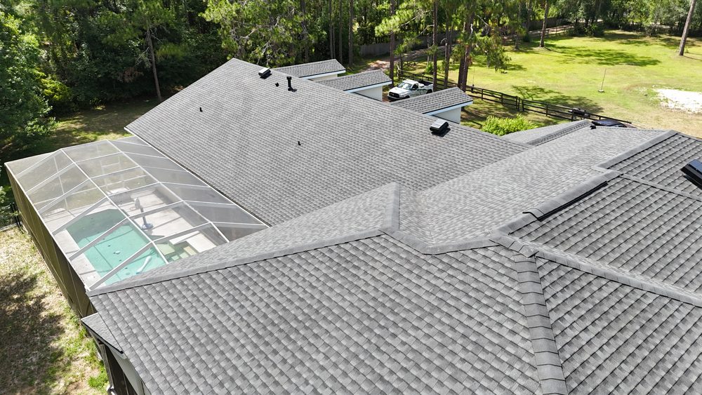 Aerial view of a large house with a gray shingle roof, multiple roof peaks, and a screened-in backyard pool area, surrounded by trees and grassy yard.