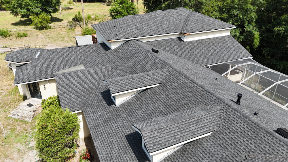 Aerial view of a house with multiple dark gray shingled roof sections, surrounded by greenery and a fenced yard. Part of a screened patio is visible on the right side.