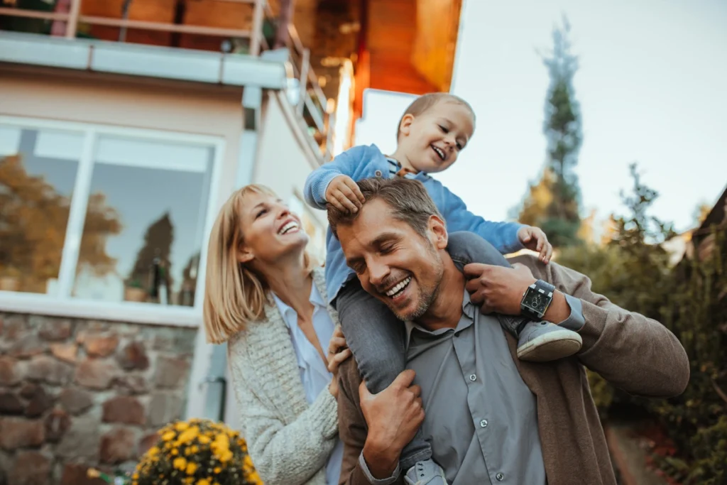 A smiling man carries a happy young child on his shoulders while a woman stands beside them, all laughing together in front of their home, proud of the new roofing Pasco County experts installed amid trees and flowers.