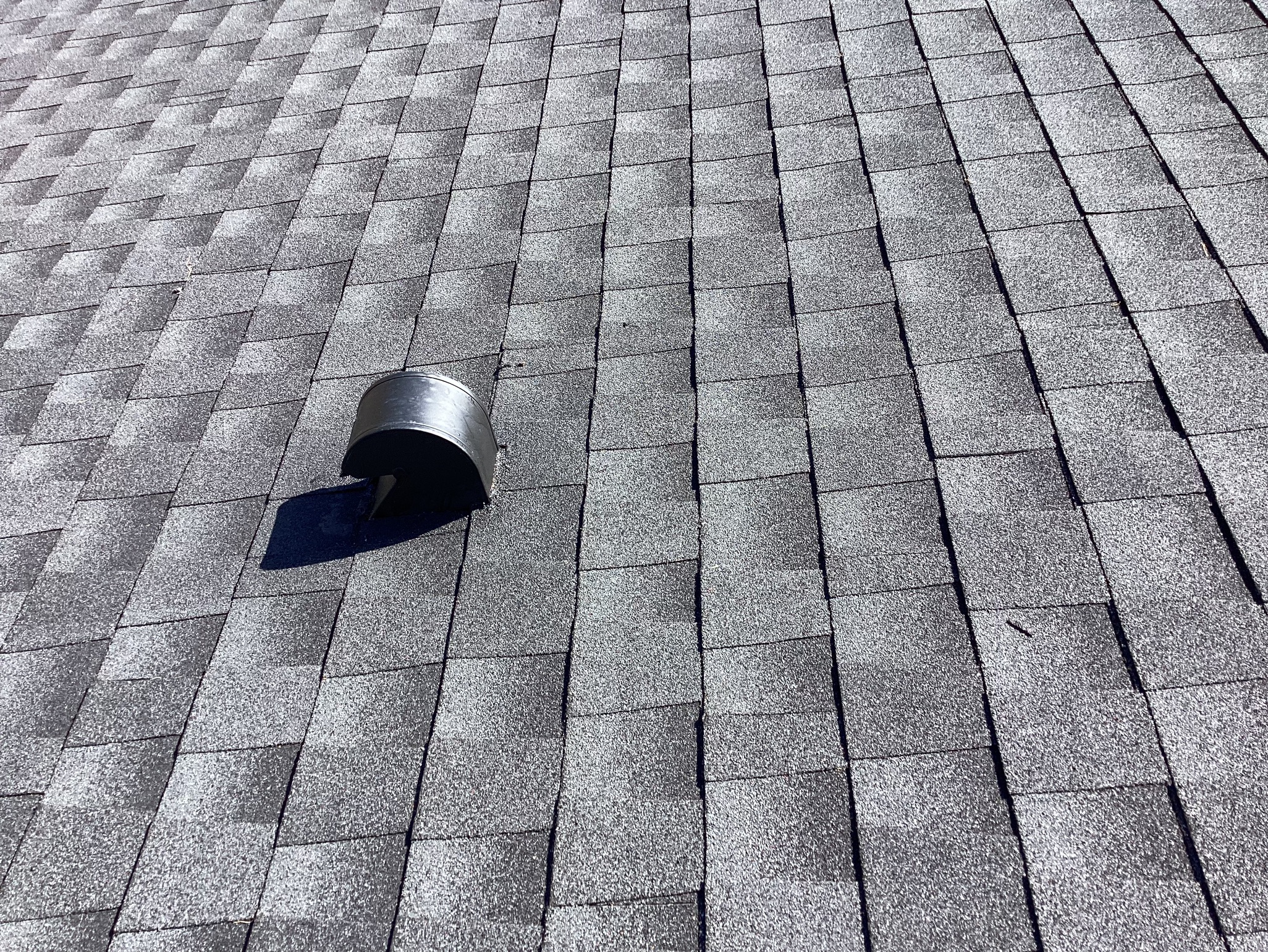 Gray asphalt shingles cover a sloped roof with a black metal vent protruding from the surface near the center left of the image. The shingles are arranged in a neat, uniform pattern.