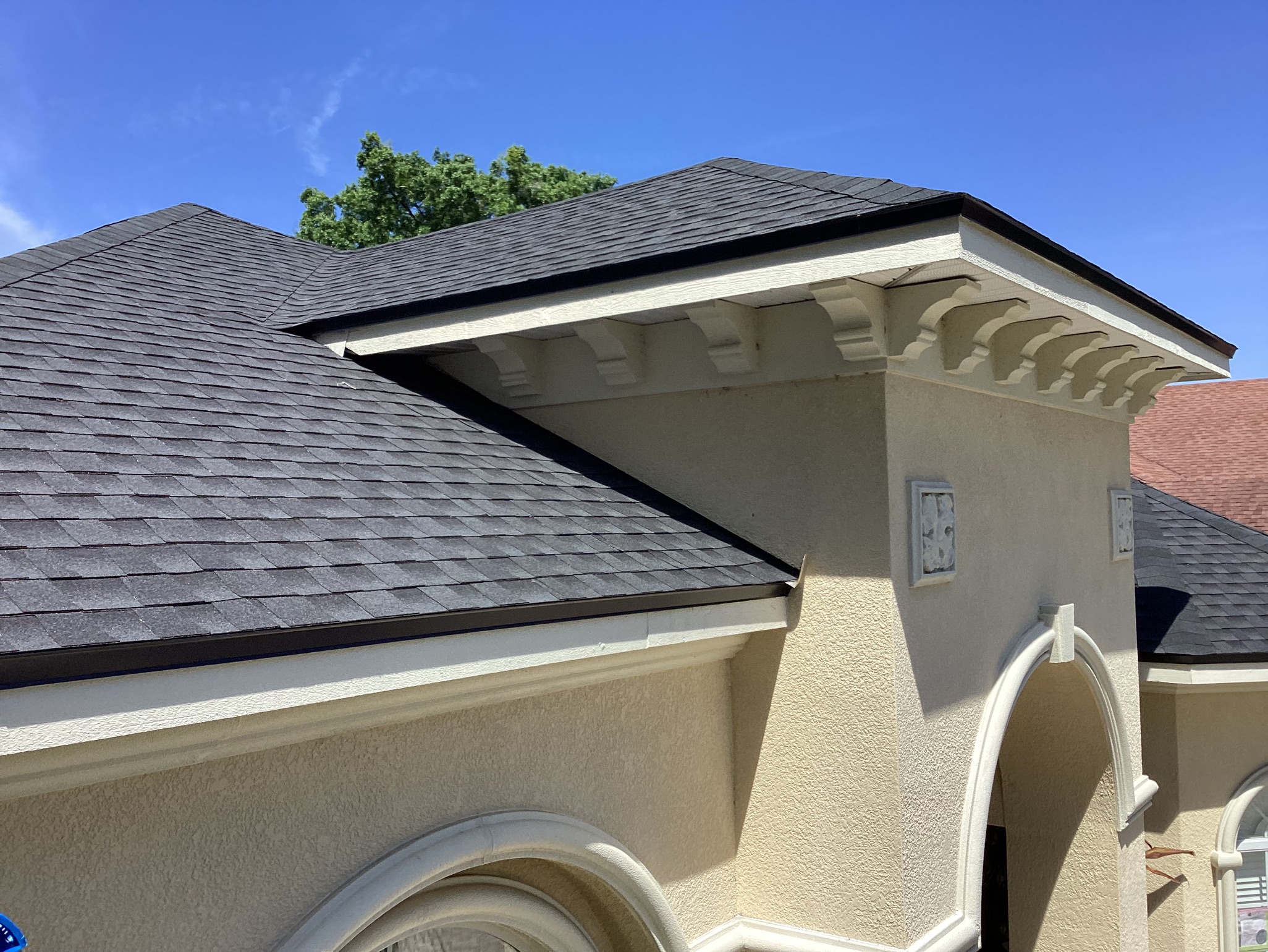 A close-up view of a house roof with dark gray shingles, beige stucco walls, decorative trim, and a bright blue sky in the background.