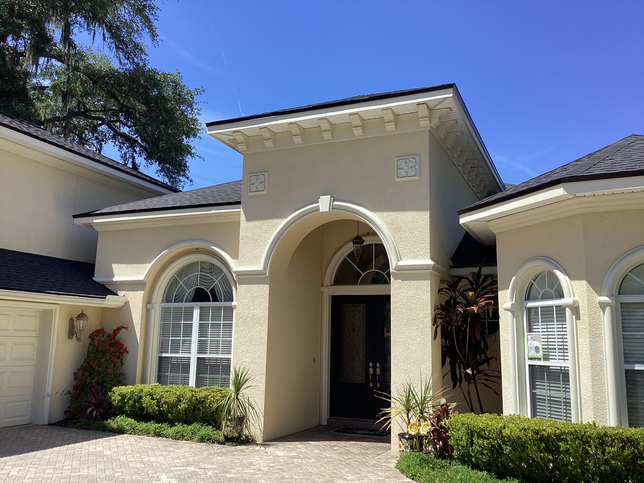 Beige, stucco house with arched windows and a double-door entry under a large archway, surrounded by green shrubs and tropical plants, under a clear blue sky.
