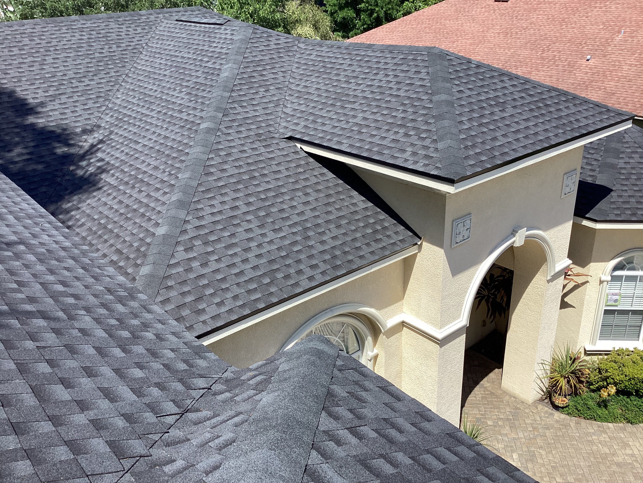 A close-up view of the dark gray shingle roof on a beige stucco house with tall arches and a paved driveway, with part of another red shingle roof visible in the background.