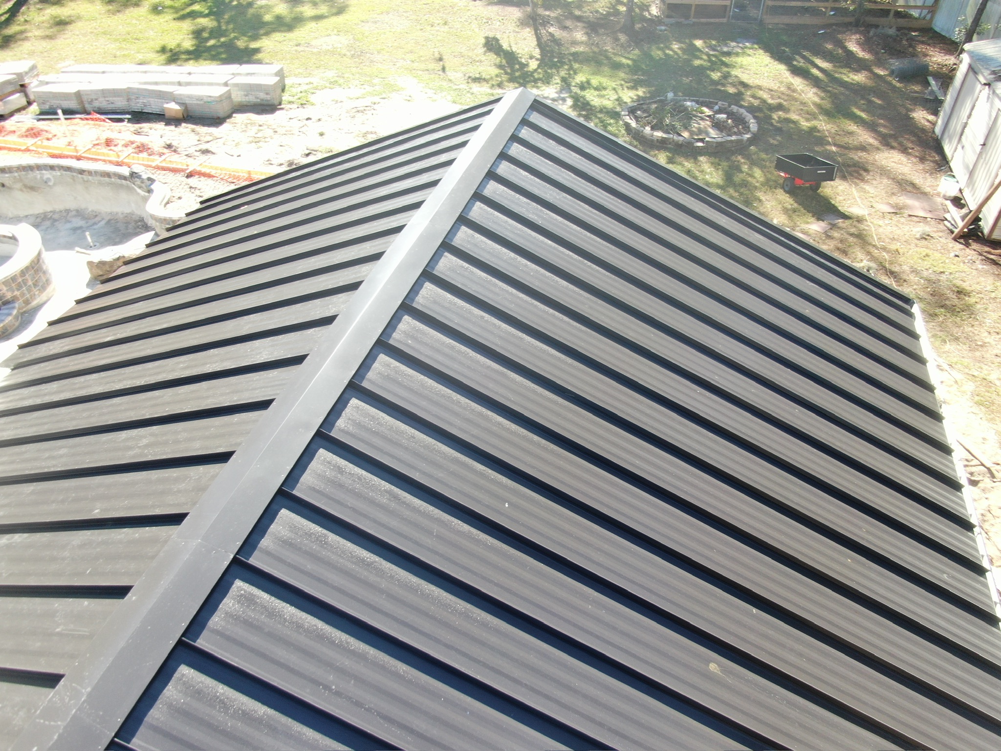 A close-up, top-down view of a black metal roof with raised seams, surrounded by a construction area with scattered building materials and patches of grass.