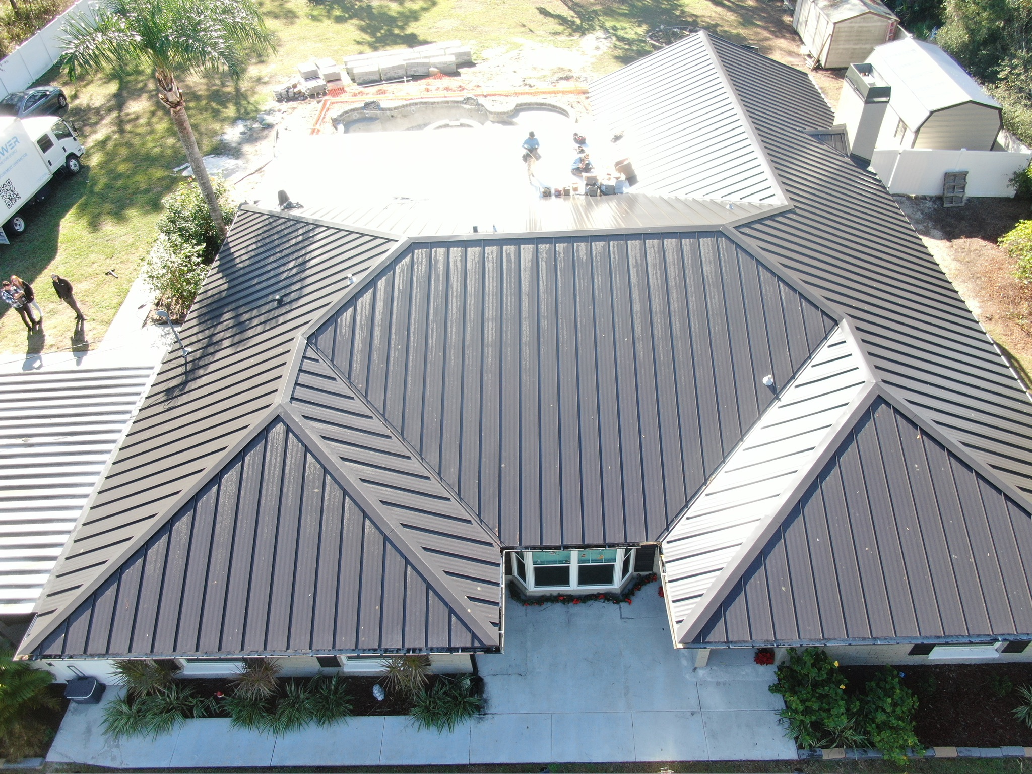 Aerial view of a house with a dark metal roof, surrounded by a driveway, yard, trees, and a swimming pool under construction in the backyard. Several people are visible near the pool and on the driveway.