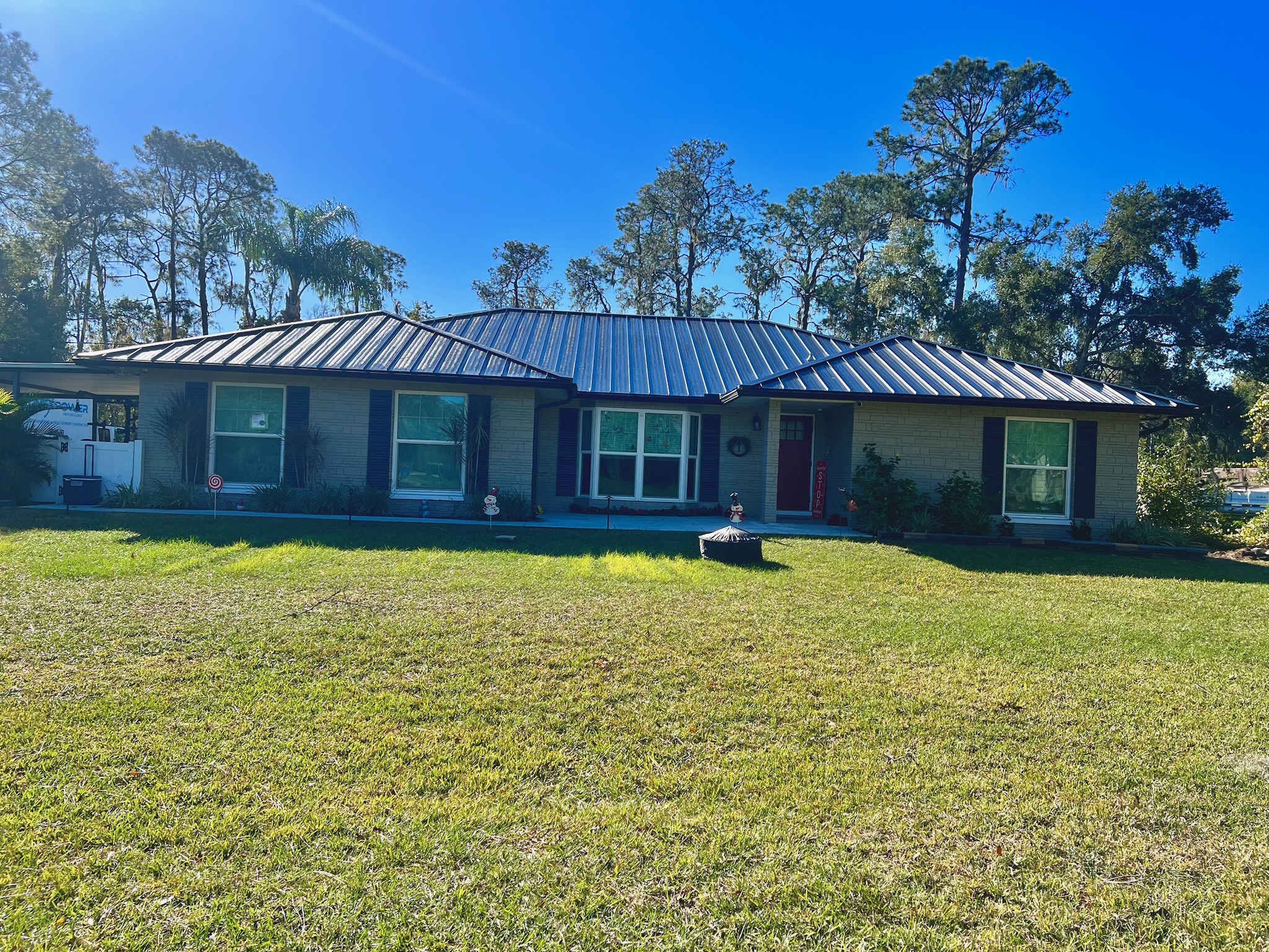 A single-story house with a metal roof, three sets of windows in front, a large grassy lawn, and trees in the background under a clear blue sky.