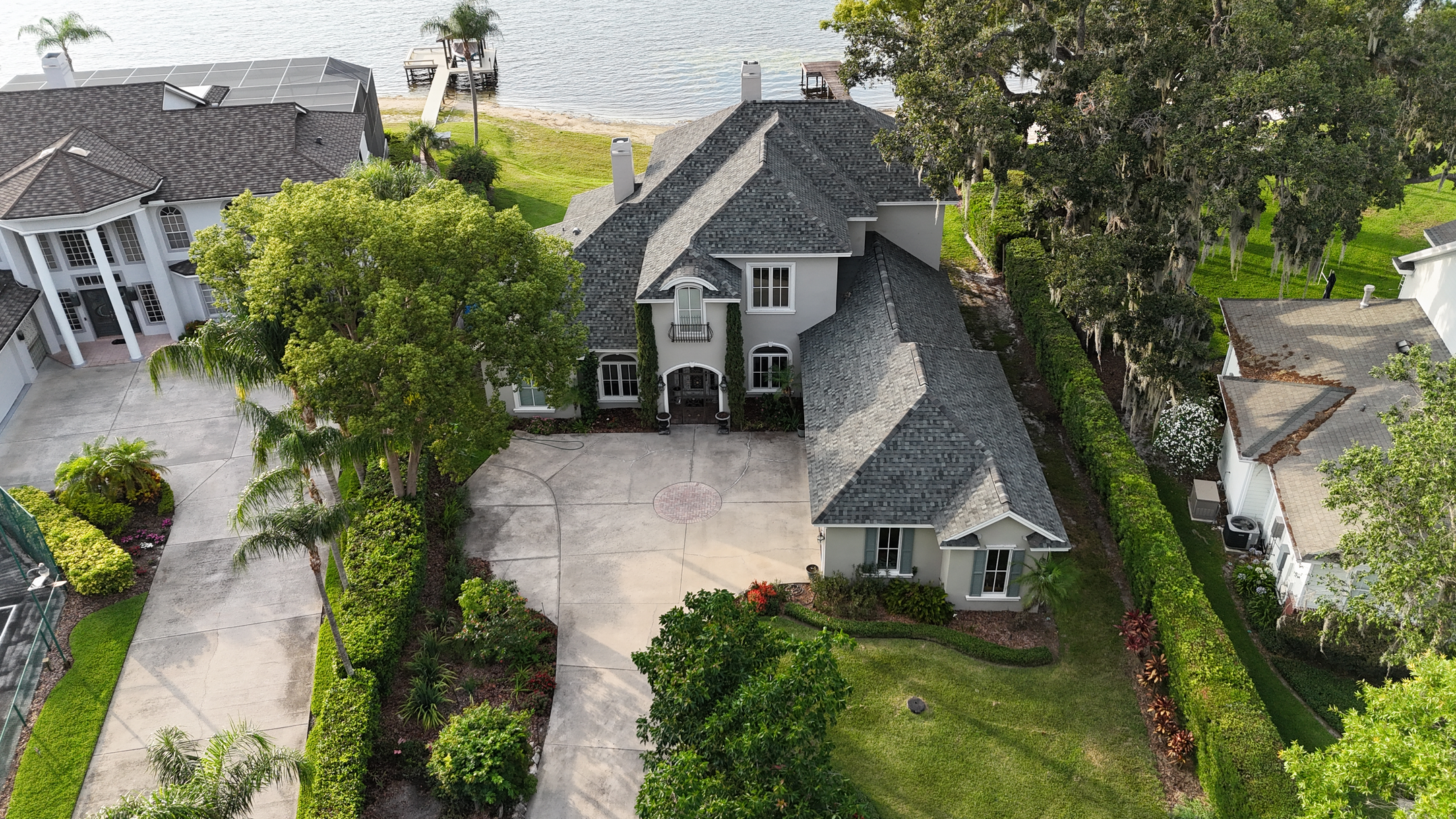 Aerial view of a large, two-story gray house with a manicured yard, surrounded by trees and greenery, located next to a lake with a dock in the background.