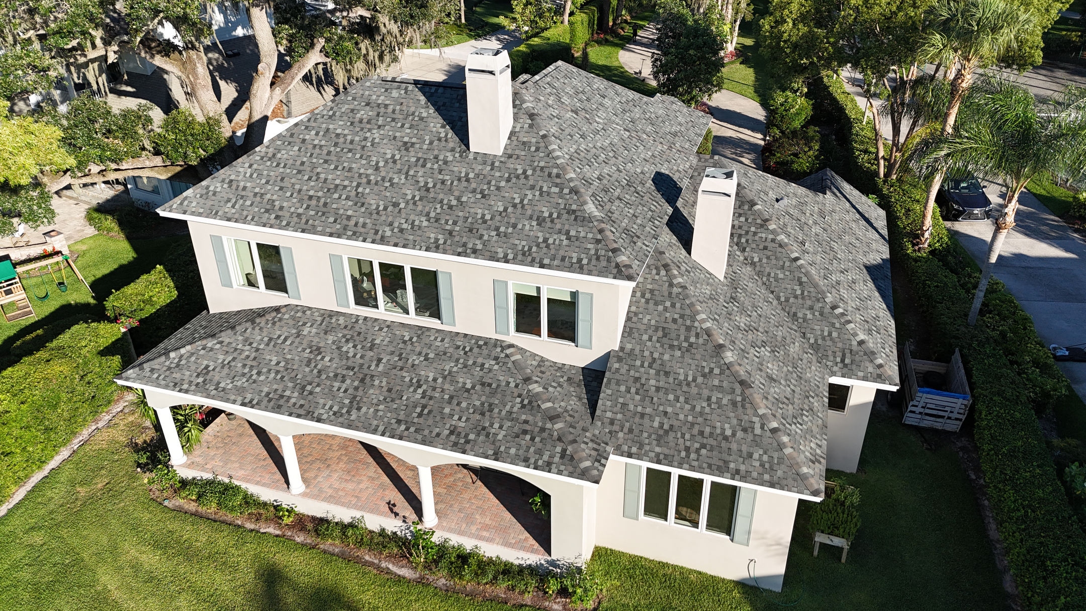 Aerial view of a two-story house with light-colored walls, white trim, and a gray shingled roof. The home features a front porch with white columns and is surrounded by green grass and trees.
