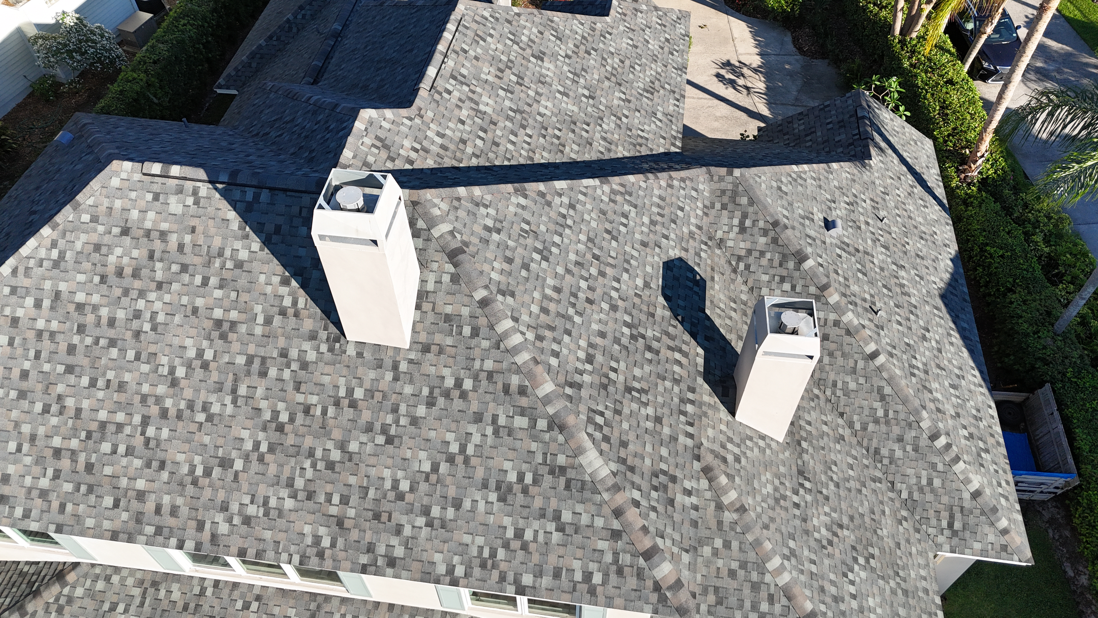 Aerial view of a house roof with multiple gables and two white chimneys, covered in gray asphalt shingles, surrounded by green trees and bushes.