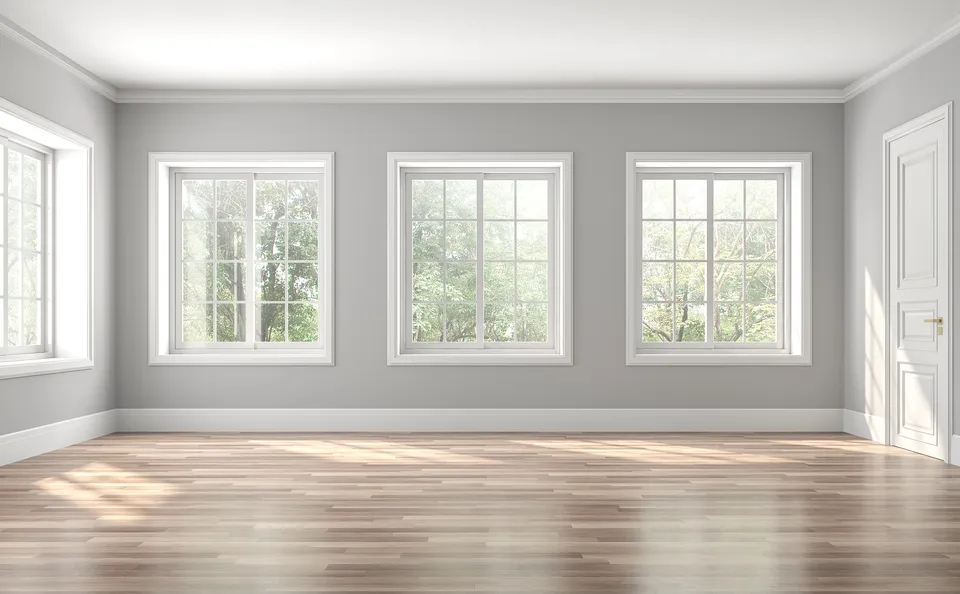 Bright, empty room with three large windows, light gray walls, white trim, and wood flooring. Sunlight streams in, and green trees are visible through the windows. A closed white door is on the right.