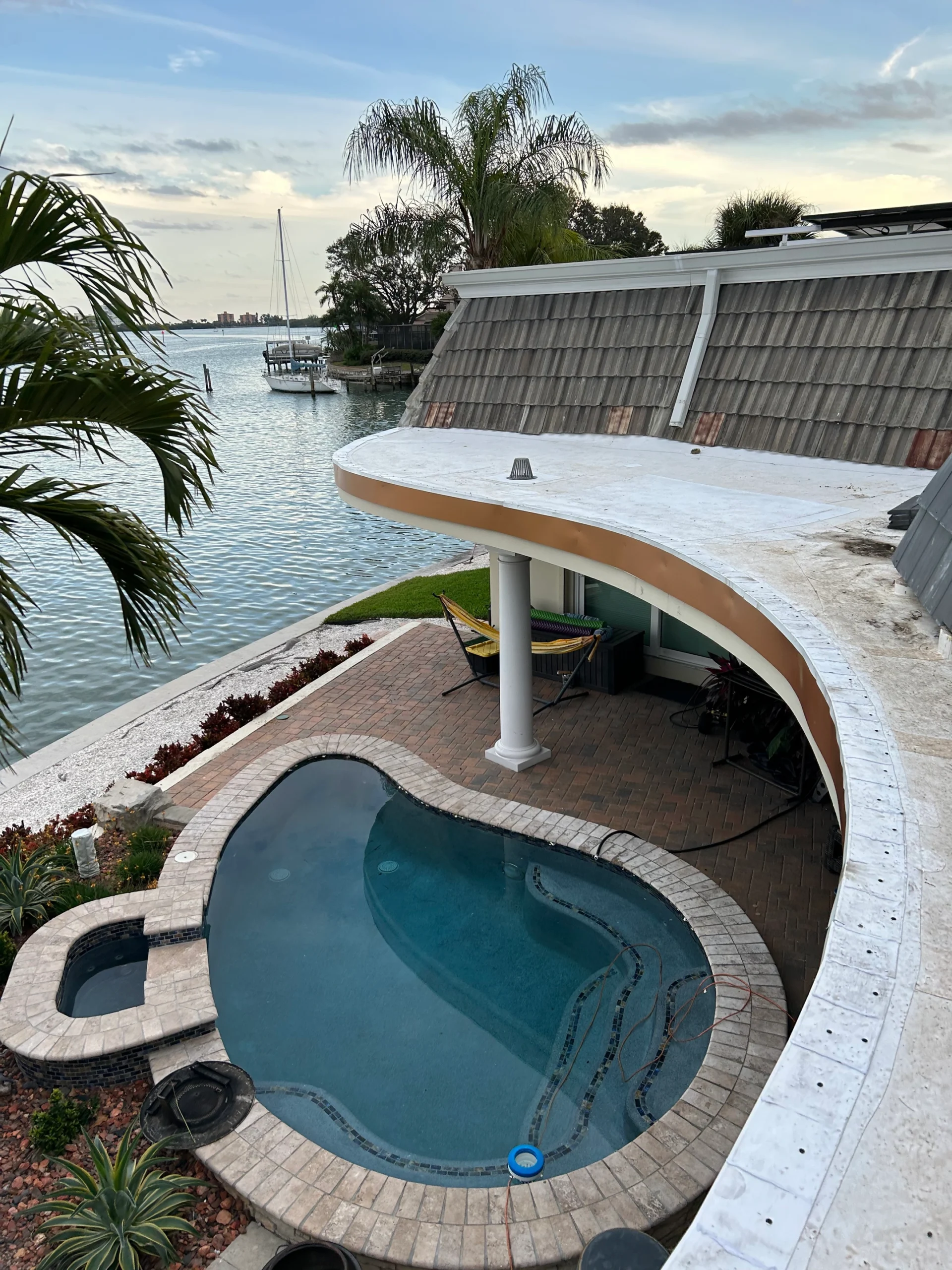 View of a small, curved swimming pool beside a waterfront patio with a hammock, palm trees, and a sailboat docked nearby; the scene is calm under a partly cloudy sky at sunset.