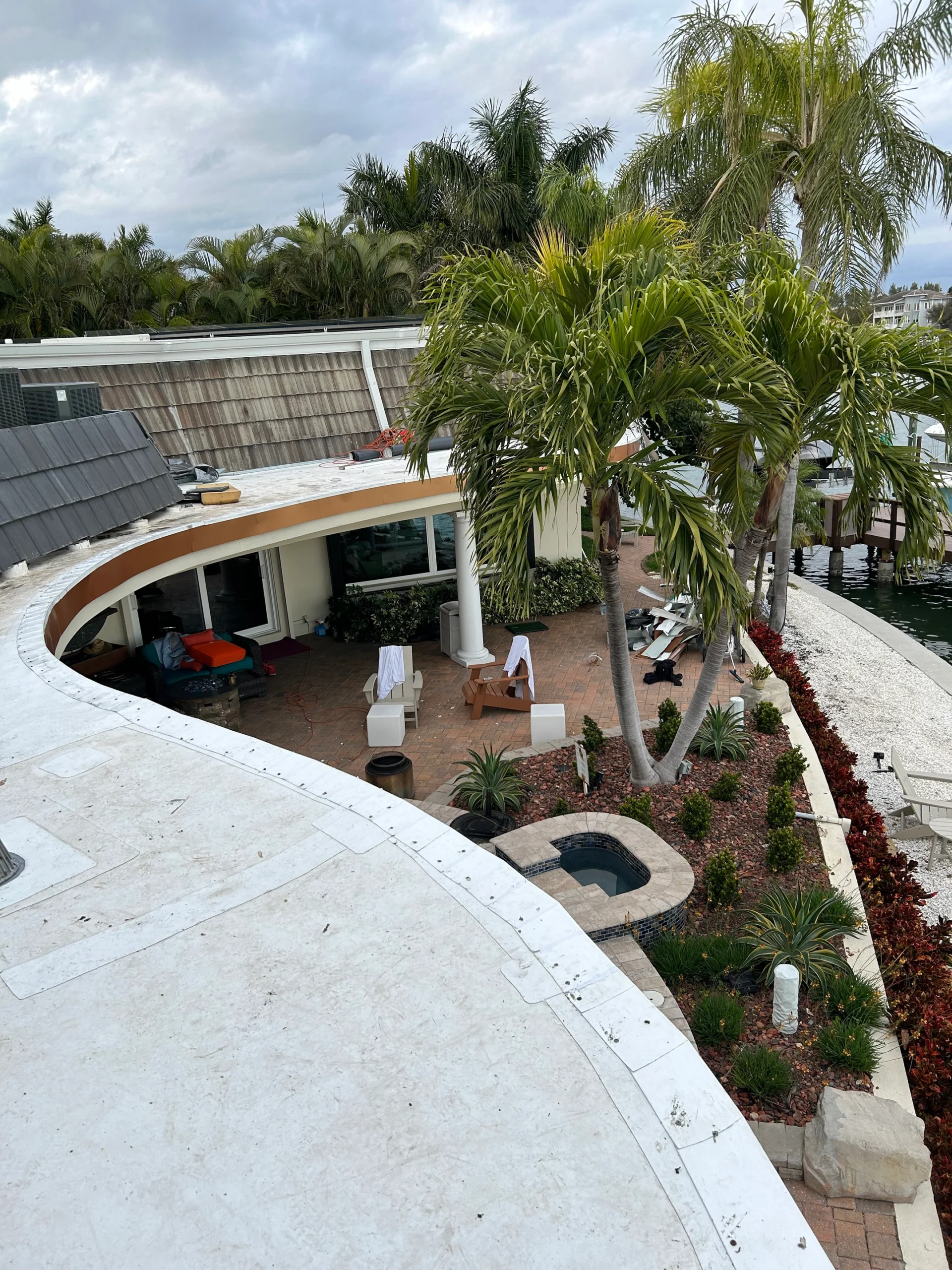 A curved white rooftop overlooks a patio with chairs, palm trees, and landscaped garden beds next to a waterfront. The sky is cloudy, and lush greenery surrounds the area.