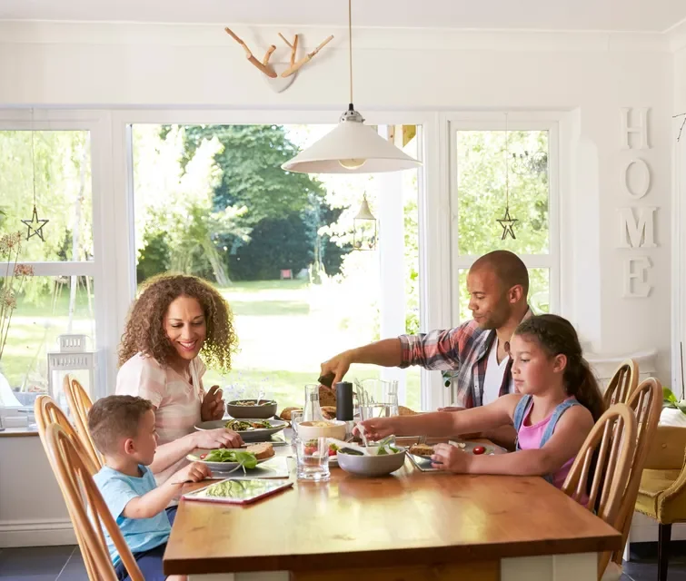 A family of four sits around a wooden dining table, enjoying a meal together in a bright, sunlit room with large windows overlooking a green garden.