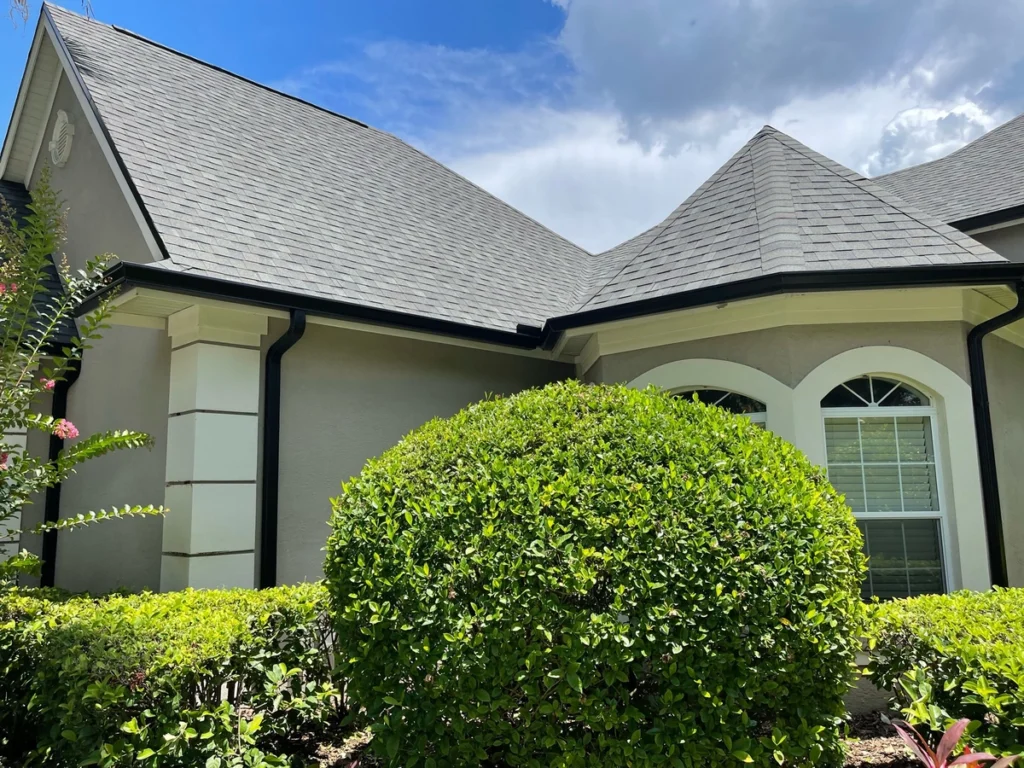 A neatly trimmed round green bush sits in front of a beige house with white trim, arched windows, and a dark gray shingled roof—a perfect example of quality roofing Pinellas County homeowners appreciate—under a partly cloudy sky.