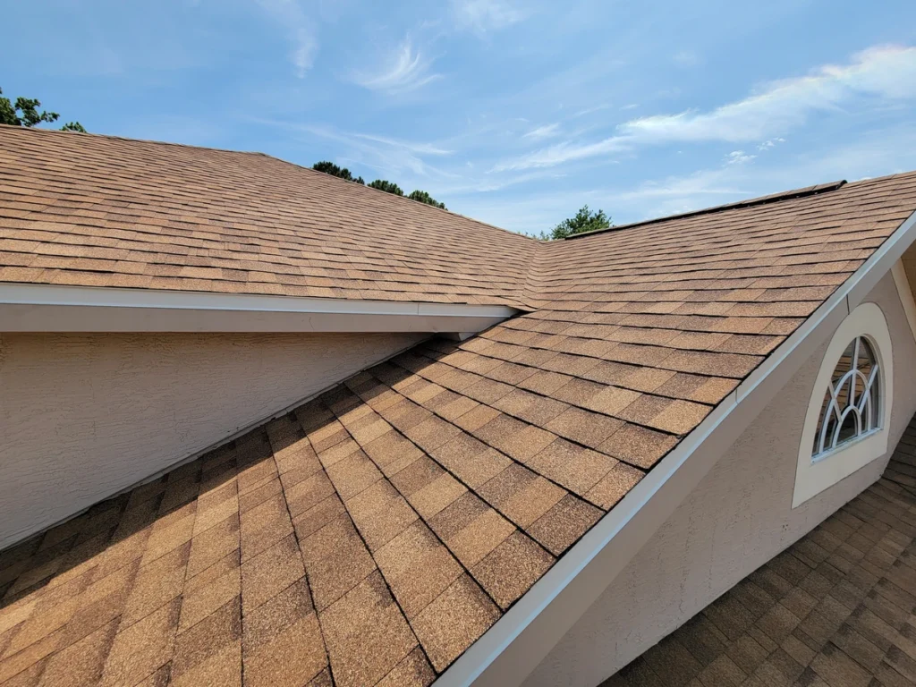 A close-up view of a house roof with brown asphalt shingles under a blue sky, showcasing intersecting rooflines and a partial arched window on the right—an example of expert roofing Riverview homeowners can trust.