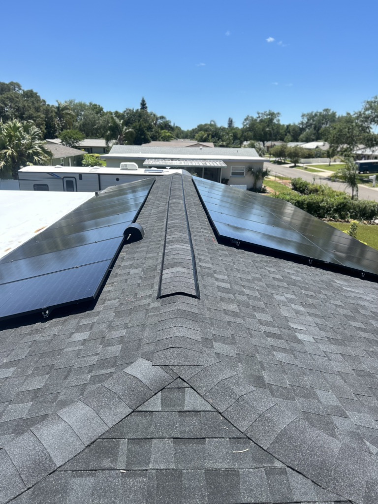 Rooftop with dark gray shingles featuring two symmetrical rows of solar panels installed on either side of the ridge, under a clear blue sky in a suburban neighborhood.