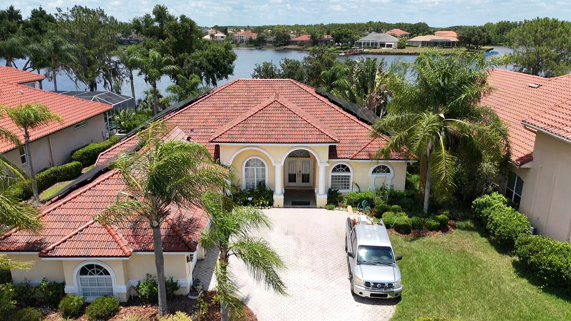 A single-story house with a red tile roof and arched windows, surrounded by palm trees and greenery. A silver pickup truck is parked in the driveway, and a lake with houses is visible in the background.