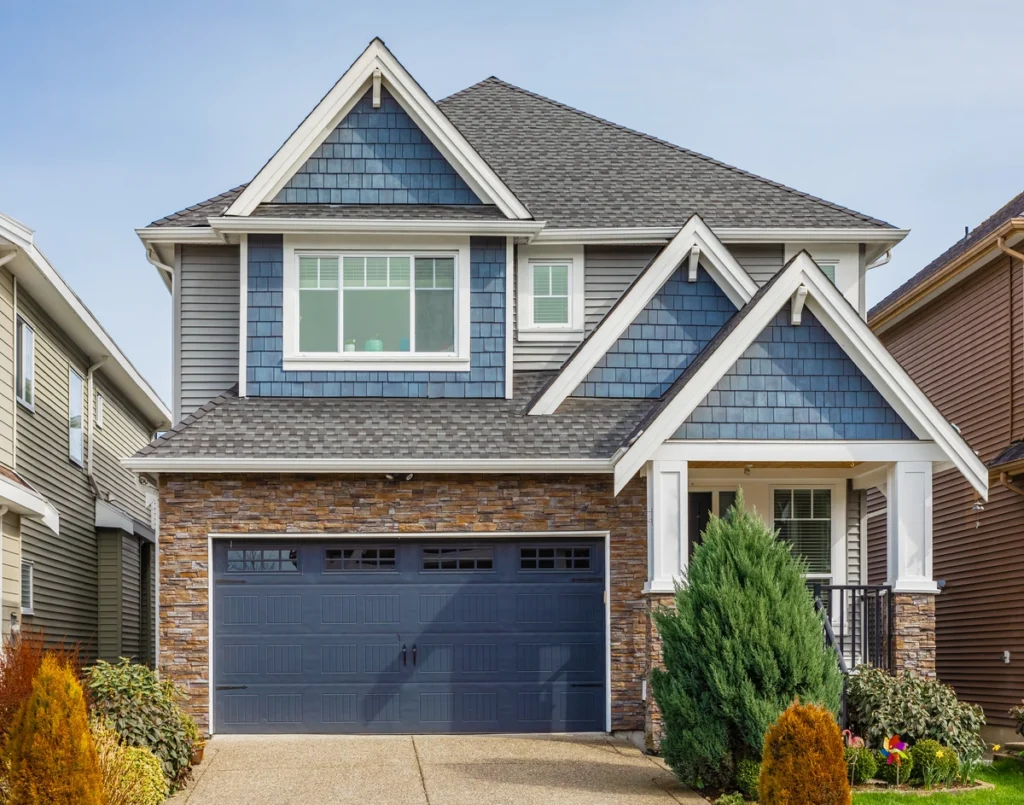 Two-story modern suburban house with blue siding, stone accents, quality roofing Lakeland craftsmanship, a double garage, and landscaped front yard with shrubs and a small porch.