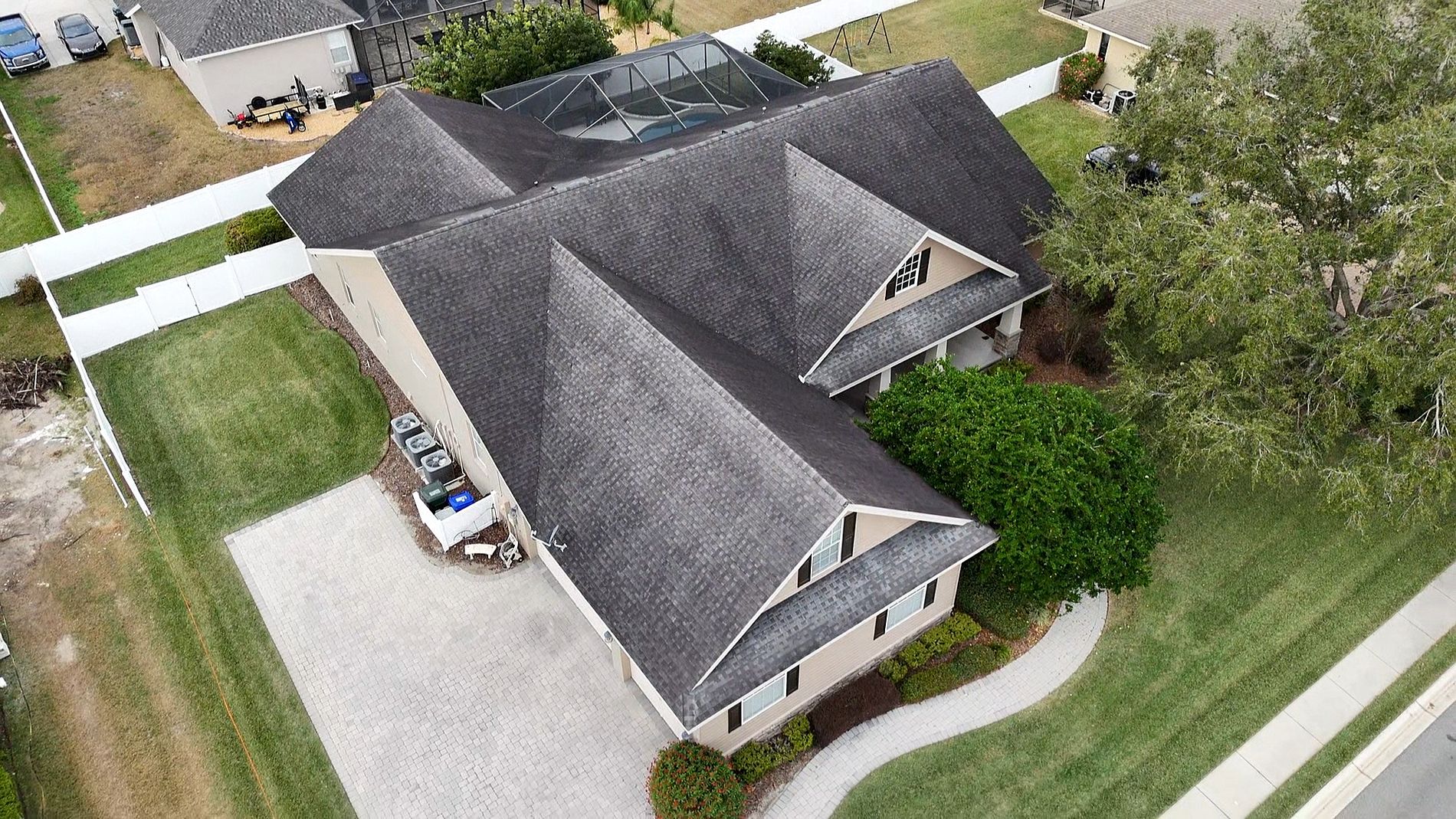 Aerial view of a large suburban house with a gray shingle roof, white trim, paved driveway, and neatly landscaped yard surrounded by a white fence. A screened pool enclosure is visible at the back of the house.