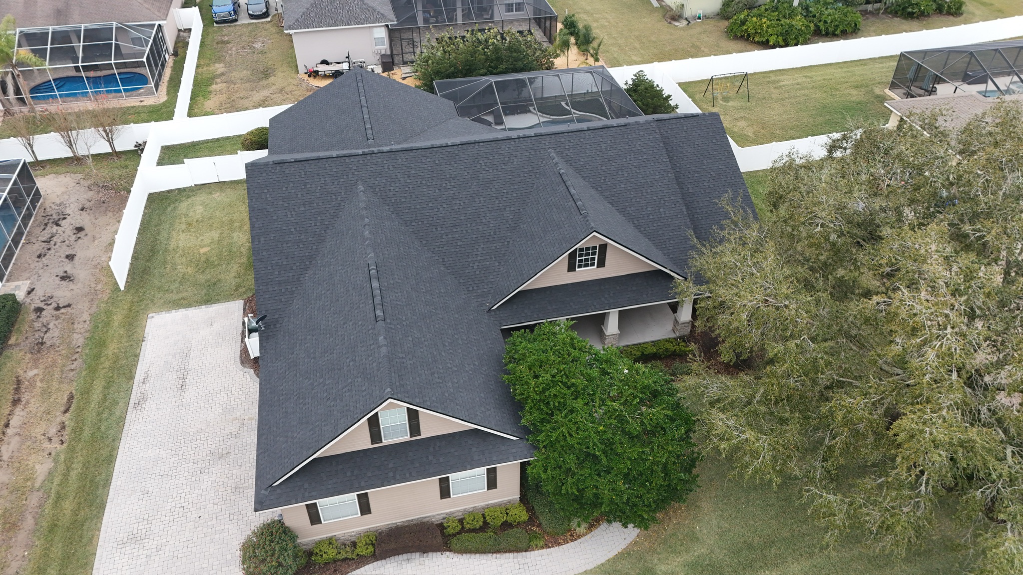 Aerial view of a beige house with a dark gray roof, surrounded by a white fence and green lawn, with a paved driveway and landscaped bushes in the front. Neighboring houses and trees are visible in the background.