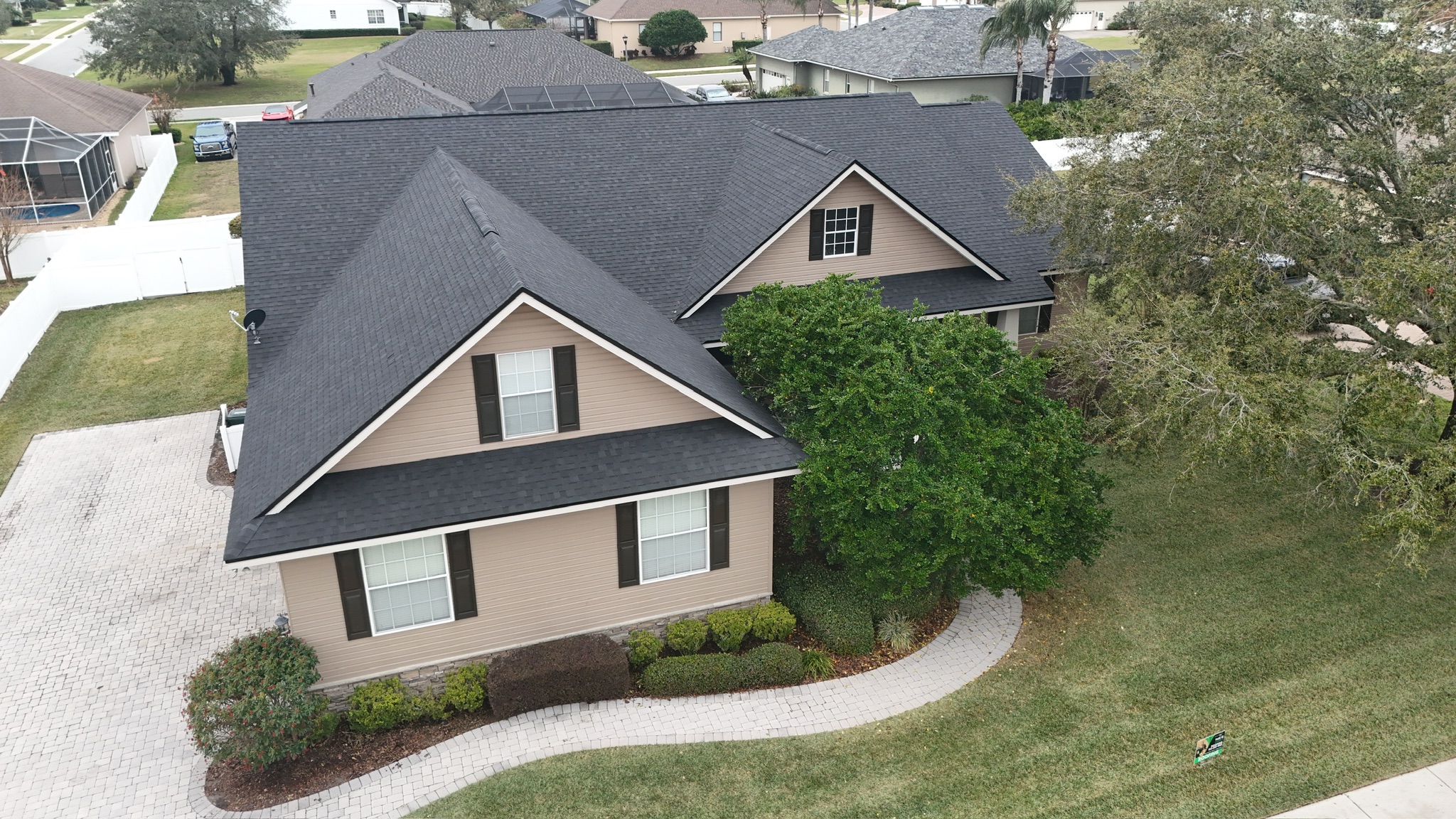 Aerial view of a tan, two-story house with a dark gray roof, white-trimmed windows, and well-kept landscaping, including green bushes and a curved walkway leading to the entrance.
