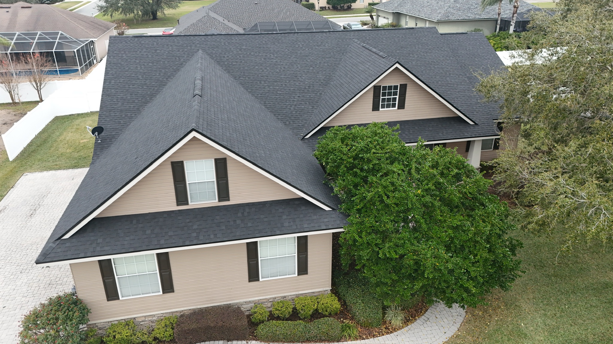 Aerial view of a tan, two-story suburban house with dark shingles, black shutters, and neatly trimmed bushes and trees in the front yard. A white fence and neighboring houses are visible in the background.